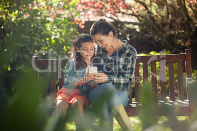 Girl showing mobile phone to mother while sitting on wooden bench