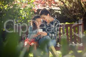 Girl showing mobile phone to mother while sitting on wooden bench