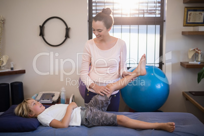 Smiling young female therapist examining knee of boy lying on bed