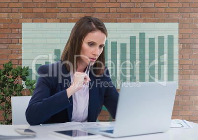Thoughtful business woman at a desk looking at a computer against brick wall with graphics