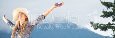 Millennial woman in sun hat with arms in air against misty mountain and tree