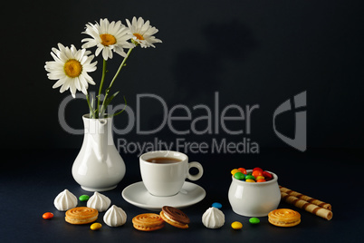 Cup with tea, teapot, sweets and white daisies on black backgrou
