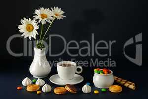 Cup with tea, teapot, sweets and white daisies on black backgrou