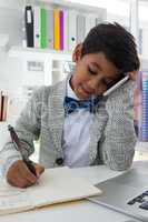 Smiling businessman writing on book while talking on smartphone