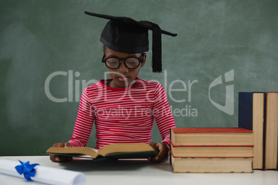 Schoolgirl in mortar board reading book against chalkboard