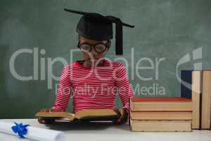 Schoolgirl in mortar board reading book against chalkboard
