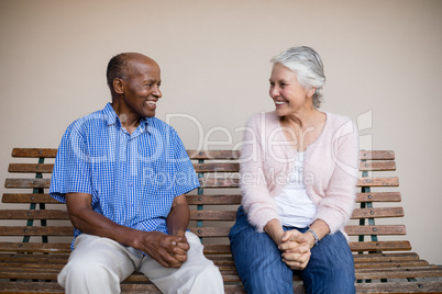 Smiling senior man and woman looking at each other while sitting on bench against wall