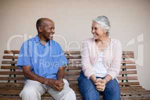 Smiling senior man and woman looking at each other while sitting on bench against wall