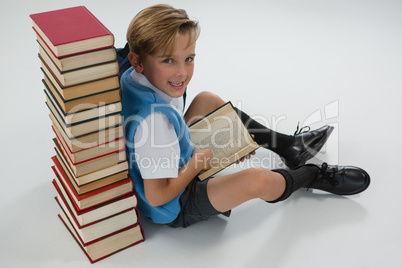 Schoolboy reading book while sitting against books stack on white background