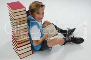 Schoolboy reading book while sitting against books stack on white background