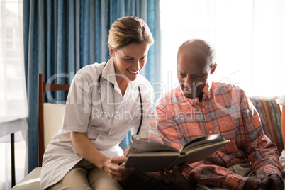 Smiling female doctor reading book with senior man sitting on furniture against window