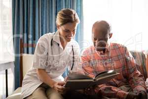 Smiling female doctor reading book with senior man sitting on furniture against window