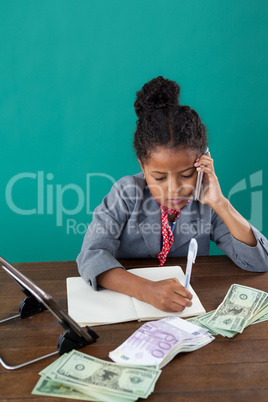 High angle view of businesswoman using phone while writing on book by paper currency