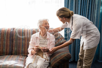 Cheerful senior woman holding knitting wool while looking at female doctor against window