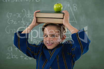 Schoolboy holding books stack with apple on head against chalkboard