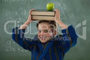 Schoolboy holding books stack with apple on head against chalkboard