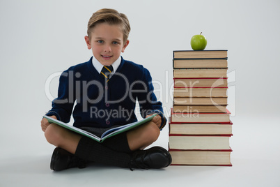 Schoolboy reading book while sitting beside books stack