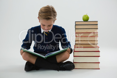 Schoolboy reading book while sitting beside books stack