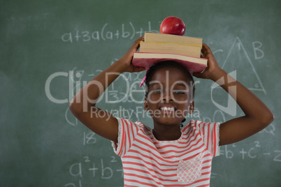 Schoolgirl holding books stack with apple on head against chalkboard