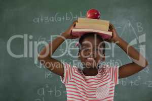 Schoolgirl holding books stack with apple on head against chalkboard