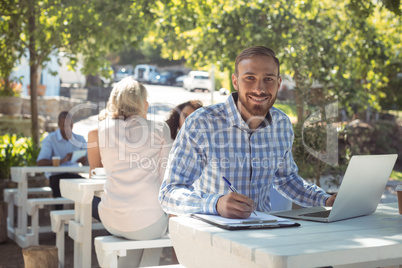 Smiling man writing on clipboard while using laptop