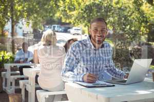 Smiling man writing on clipboard while using laptop