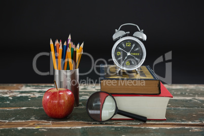 Alarm clock on stack of books with pen holder, apple, and magnifying glass