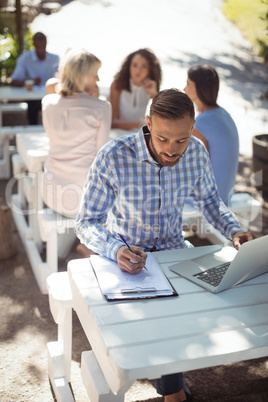 Man writing on clipboard while using laptop