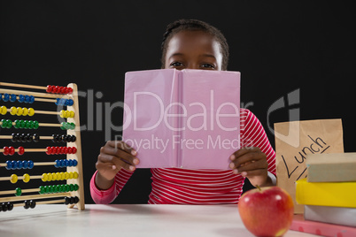 Schoolgirl hiding face behind book against black background