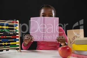 Schoolgirl hiding face behind book against black background