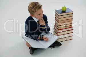 Schoolboy doing his homework while sitting beside books stack