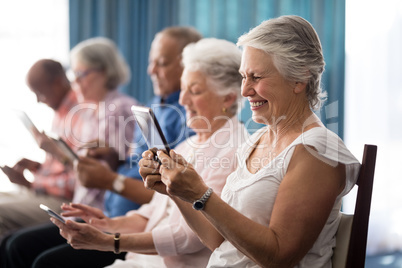 Row of smiling senior people sitting on chairs using digital tablets