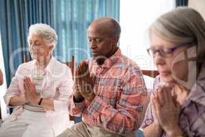 Senior man amidst women praying while sitting on chairs