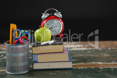 Alarm clock, apple and chalk on stack of books with pen holder