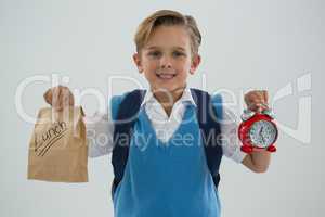 Smiling schoolboy holding alarm clock and lunch paper bag