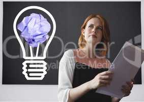 Woman holding tablet standing next to light bulb with crumpled paper ball in front of blackboard