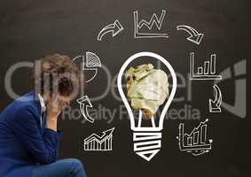 Woman sitting next to light bulb with crumpled paper ball and business graphics in front of blackboa