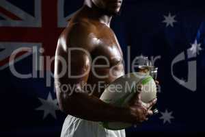 Mid section of shirtless man with trophy and rugby ball against Australian flag