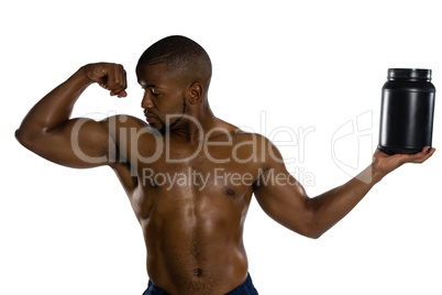 Shirtless male athlete looking at muscles while holding supplement jar