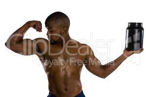 Shirtless male athlete looking at muscles while holding supplement jar