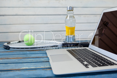 Close up of laptop by tennis racket and balls with water bottle against white wall
