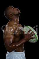 Side view of shirtless male athlete looking up holding rugby ball