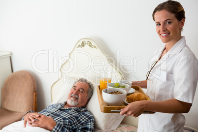 Female doctor serving food to senior patient relaxing on bed in retirement home
