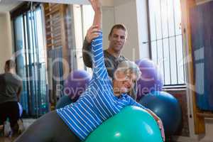 Physiotherapist assisting senior woman in performing exercise on fitness ball