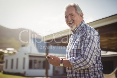 Smiling senior man with his mobile phone standing outside his house