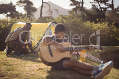 Boy playing guitar while father setting up a tent in park