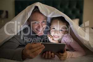 Father and daughter using digital tablet under blanket on bed