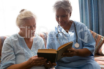 Doctor with senior woman reading book while sitting on sofa