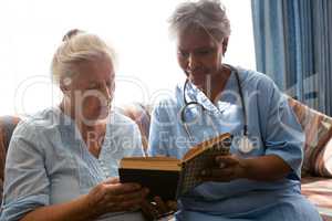 Doctor with senior woman reading book while sitting on sofa