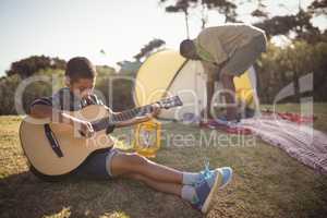 Boy playing guitar while father setting up a tent in background